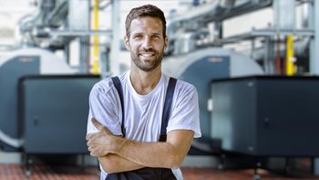 Ein lächelnder Mann in Arbeitskleidung steht mit verschränkten Armen vor großen Maschinen in einer industriellen Umgebung.